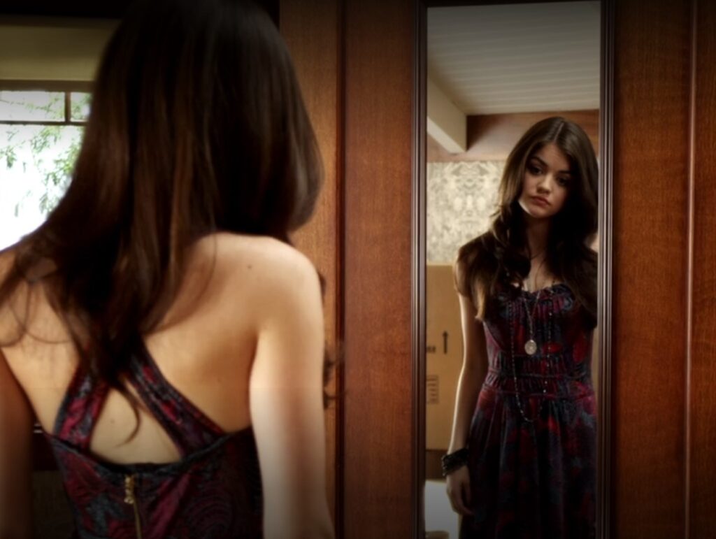 Aria is looking in the mirror, wearing a black, red and purple dress with a long necklace.