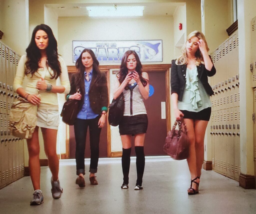 Shot of the four Liars walking down the school hallways together. Hanna is wearing a pale green ruffly top with a blazer over it, black shorts, and black strappy platform heels