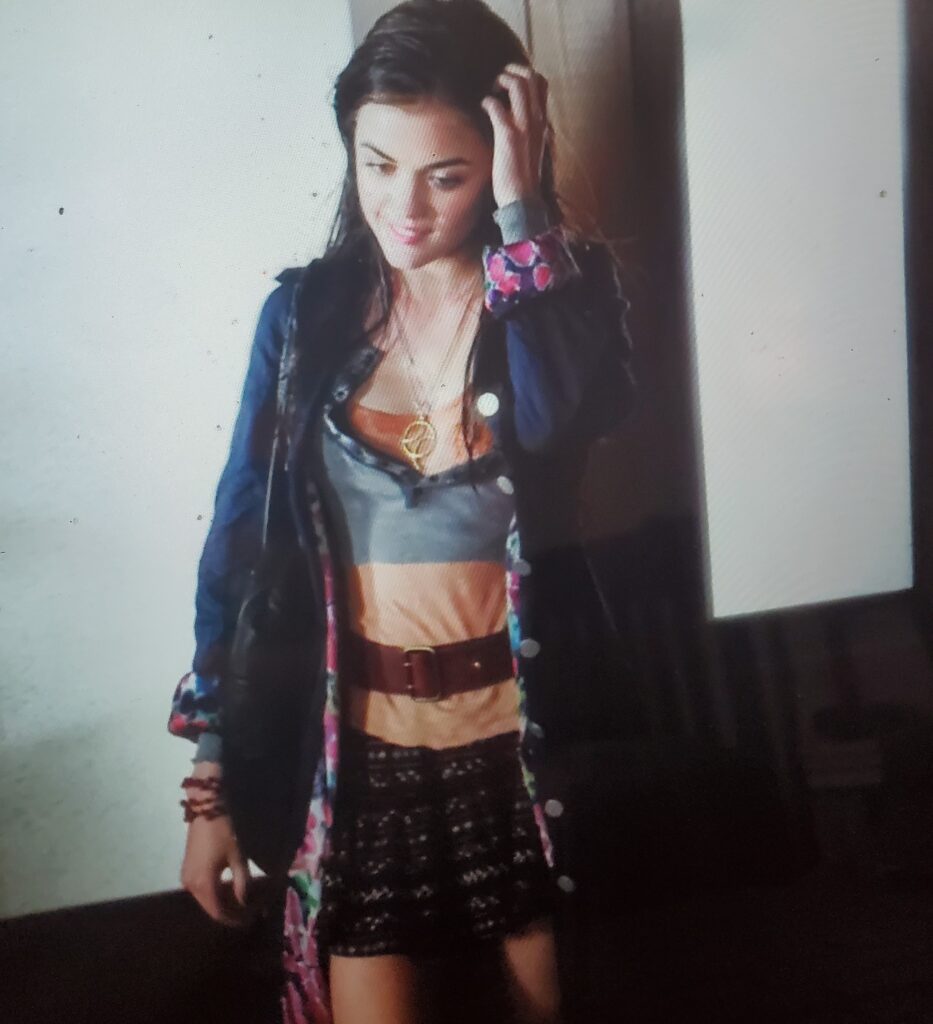 Aria wearing a gray and orange shirt with a weird layering effect, covered by a brown belt, and a knitted skirt, all covered with a knee-length jacket that is blue on the outside and brightly patterned on the inside, along with a necklace with a gold geometric pendant. She is soaked through and clearly just come out of the rain.