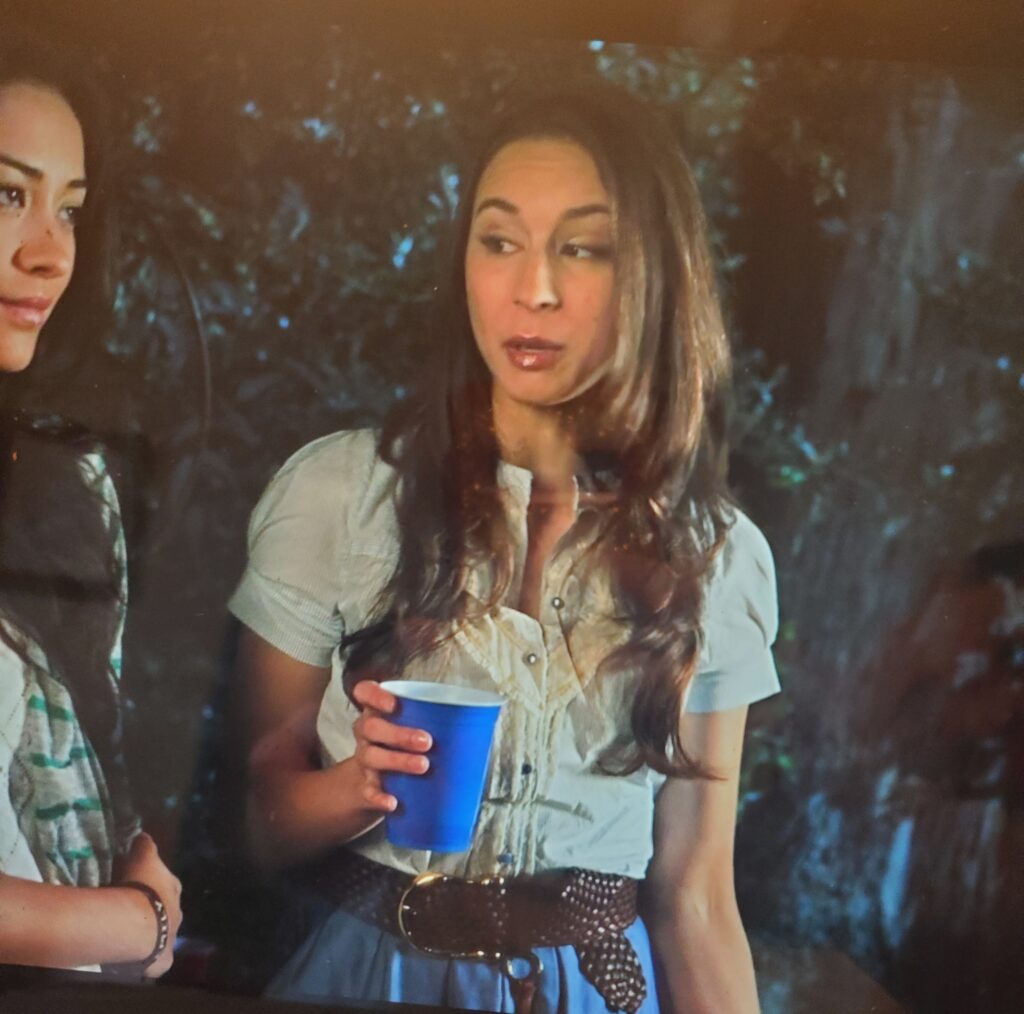 Shot of Spencer standing next to Emily and holding a blue Solo cup. She is wearing a ruffly white top with a blue skirt and a brown belt