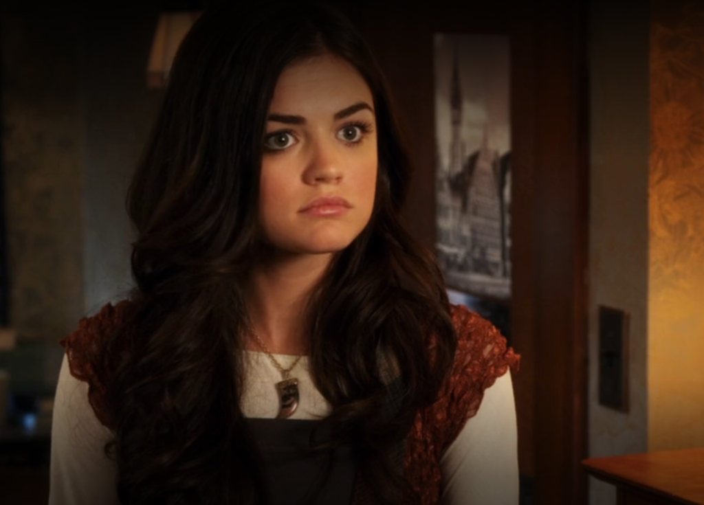 Close-up shot of Aria's upper body, where you can see that along with the white long-sleeve shirt and fuzzy dress on top of it, she is also wearing a necklace with a pendant that looks like an animal's tooth covered in gold enamel.