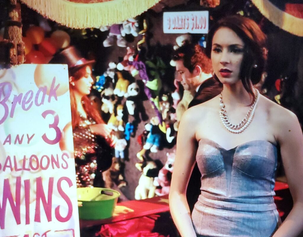 Spencer standing at a darts game stall, wearing her strapless silver dress and weird necklaces, with her hair in an updo that looks okay