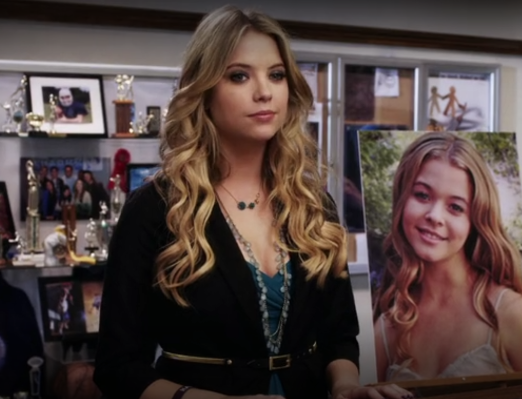 Hanna standing at a podium at Ali’s memorial service. We can see Hanna from the waist up, and she is wearing a blue dress with a black belted blazer over it and two necklaces, one long and one short.