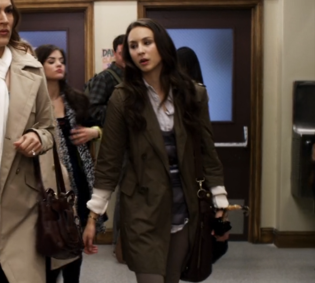Wide shot of Spencer walking in the school hallway next to her mother. She is wearing a knee-length greenish-brown trenchcoat-like jacket with large black buttons, over a white button-down shirt and a sweater-vest.