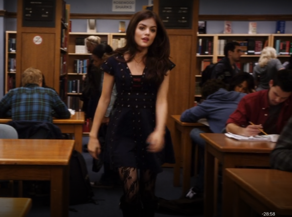 Wider shot of Aria walking in the library, where we can see that she is also wearing black tights patterned with flowers.