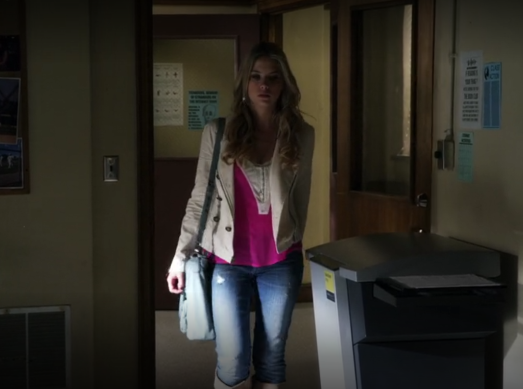 Wide shot of Hanna walking into the yearbook room; she is wearing jeans with a pink and white top (or possible two tops layered) with a white jacket.