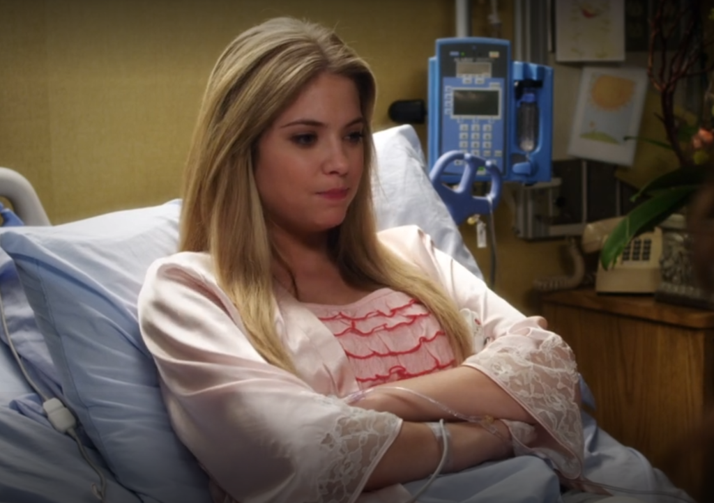 Hanna sitting up in a hospital bed, her arms folded over her chest, wearing pink silk pajamas with lacy edges on the sleeves and ruffles with a darker pink trim across the front