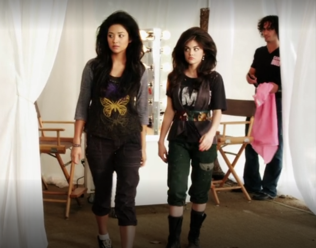 Wide shot of Aria and Emily coming out of the hair salon at Camp Mona. Both of them have had blow-outs that make their hair look huge. Emily is wearing a three-quarter sleeve top with gray sleeves, a black torso, and large butterflies embroidered on it in purple, yellow, and gray, with dark gray capri cargo pants and sneakers. Aria is wearing a black graphic t-shirt covered with a gray vest and a wide waist belt over the vest that appears to have giraffes on it, with green capri pants and calf-length boots.