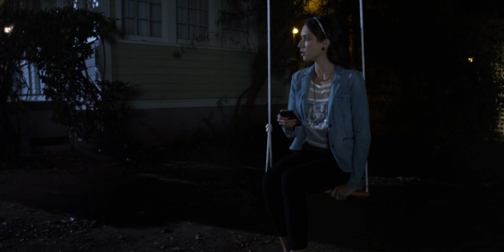 Spencer sitting on a tree swing outside in the dark, holding her phone and looking onto the side. She is wearing three-quarter length black pants, a white lacy blouse, and a light blue blazer, with a beaded white headband
