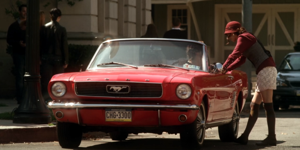 Spencer talking to Alex, who is in his red Mustang convertible in the middle of Rosewood. Spencer is wearing a felt red hat, a matching red button-down with a gray vest over it, a white lacy skirt, and knee-high socks with short boots.