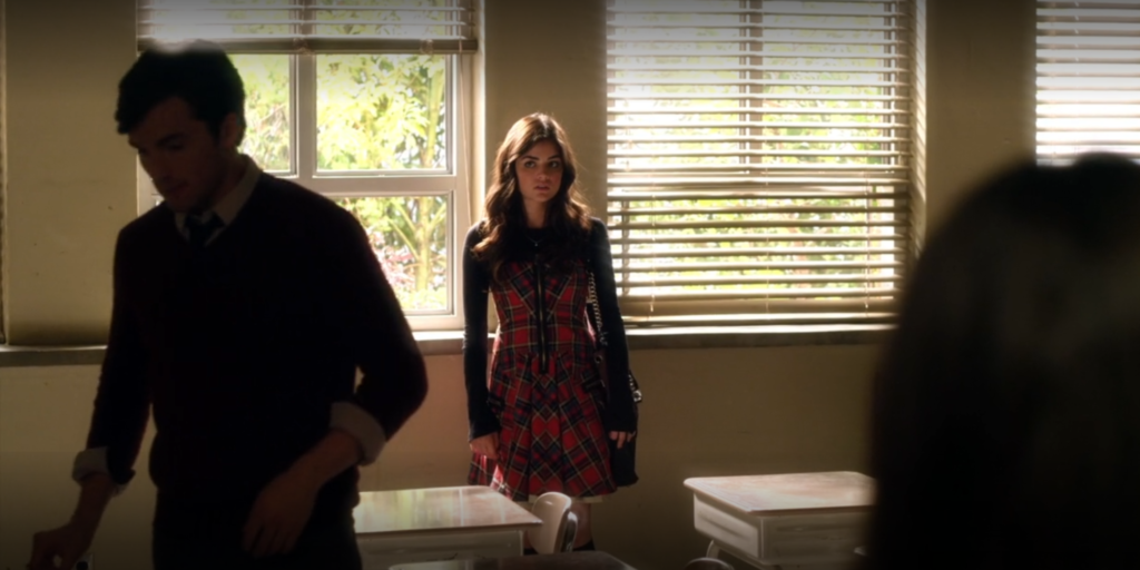 Aria in Ezra’s classroom, standing by the window lookup upset while he gets something at his desk. Aria is wearing a red and black plaid dress without sleeves over a black long-sleeved top.