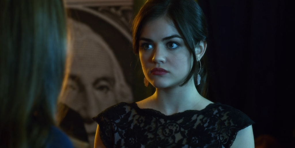 Close-up shot of Aria looking annoyed. We can see just the shoulders of the black lacy short-sleeved dress that she is wearing. Her hair is in a ponytail with some strands artfully arranged around her face. She is wearing red lipstick and dangly earrings with some kind of quartz-like stone on them