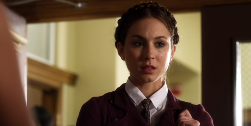 Close-up of Spencer from the shoulders up. She is wearing a black necktie with gold designs and a white button-down shirt, both of which are covered by a maroon peacoat. Her hair is all up in a braid crown.