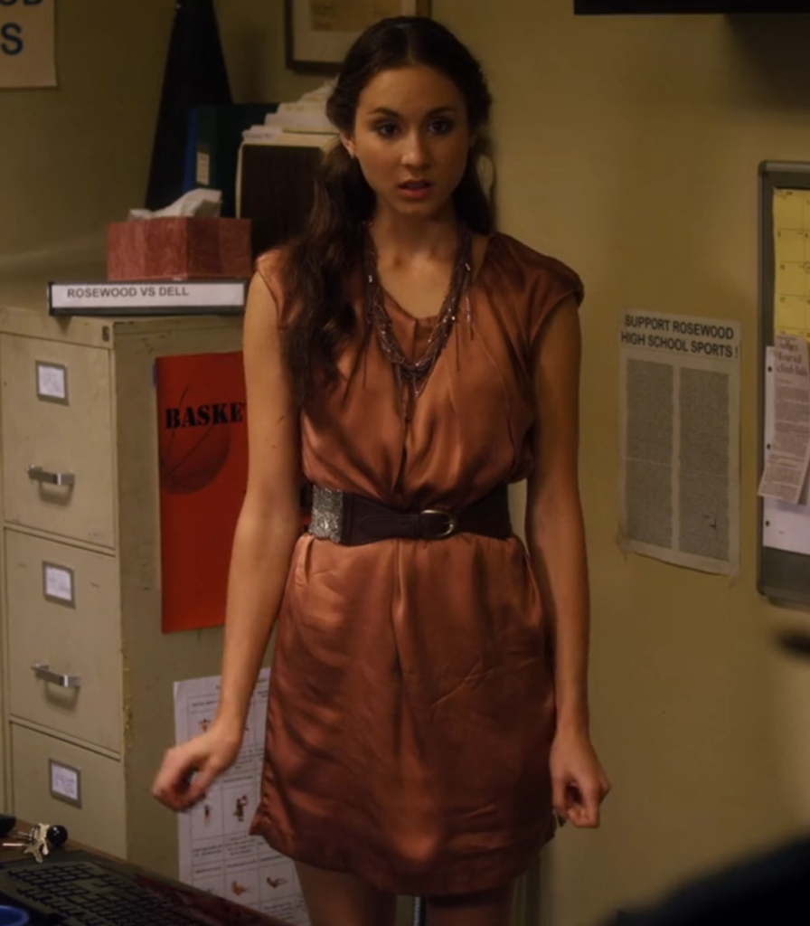 Spencer is standing in the field hockey coach’s office, looking surprised and upset, and we can see her from the knees up. She is wearing a light brown loose dress with no sleeves, which is fastened at the waist with a dark brown belt. She’s also wearing a brown thickly-beaded necklace, and her hair is half up.