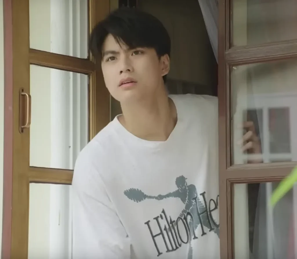 Pat from the GMMTV show แค่เพื่อนครับเพื่อน (Bad Buddy), leaning out a window and looking concerned. He is wearing white t-shirt with a large gray logo that is the silhouette of a person playing racquetball, with the words Hilton Head over it.