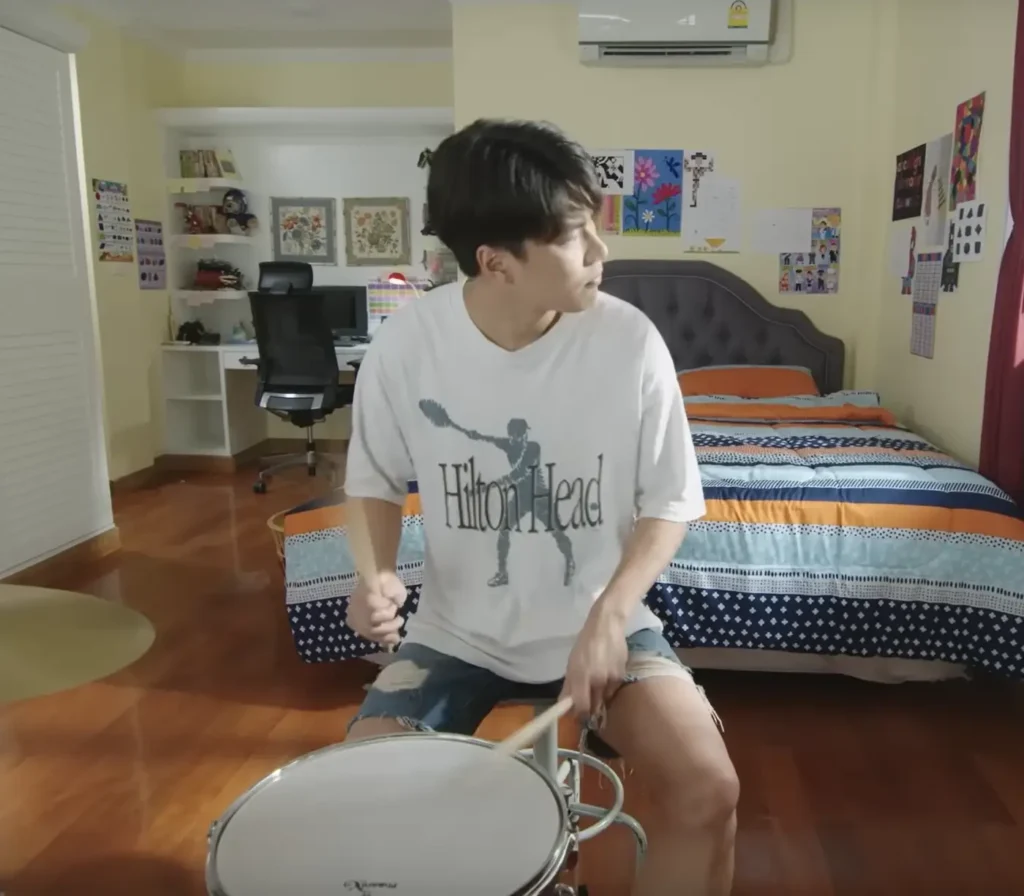 Pat wearing the same shirt, but this time sitting in his bedroom at his drum kit, playing drums and looking out the window.