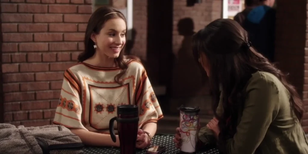 Spencer and Emily are sitting outside at a picnic bench at their school, Emily facing away from the camera. Spencer is smiling and wearing a knitted top that looks like a poncho; it is beige with geometric shapes in orange and black, including three black suns across the center of her chest. She is also wearing earrings and a headband.