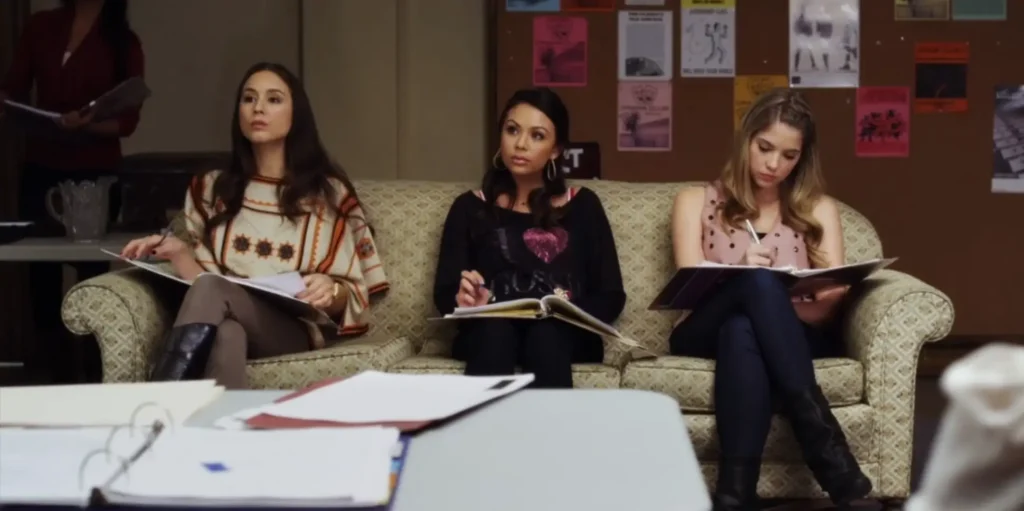 Spencer, Mona, and Hanna are all sitting on a couch at play rehearsals, each of them with a binder on their laps with their scripts. Spencer is wearing the knitted poncho but now she can see is also wearing brown pants and tall black leather boots. Mona is wearing black wide-collared sweater with a pink shiny heart on it. Hanna has her braided hair, but now we can also see that she is wearing a pale pink sleeveless top with little metal studs dotting it, with dark jeans and black leather boots.