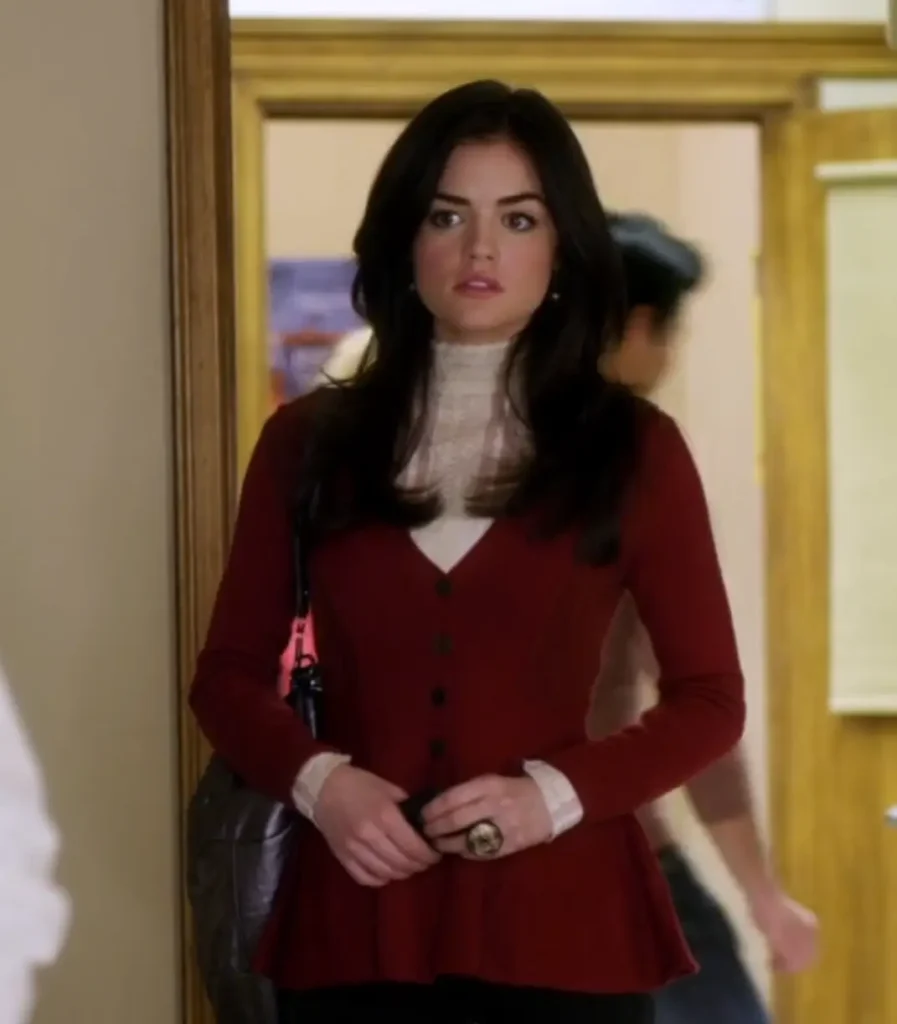 Aria is standing in the doorway of a classroom at Rosewood High, looking alarmed. She is wearing a white lacy top that goes up to her neck like a half-turtleneck, and over that she is wearing a read cardigan or blazer that she is wearing buttoned, which goes down pas her waist. She is also wearing a very large ring on one of her fingers that looks like a gold and black circular stone.