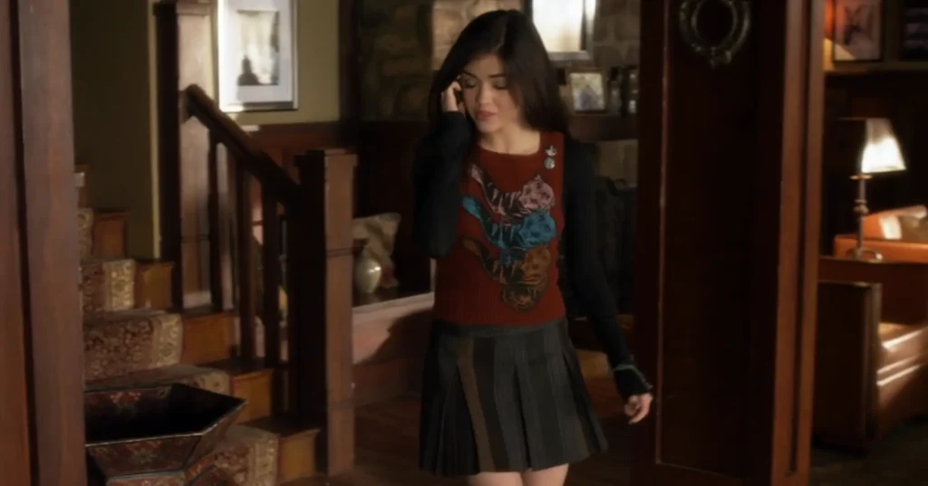 Aria is wearing a gray and brown thigh-length pleated skirt and a long sleeve dark top with a red knitted vest over it. The vest has a design with three stylized tigers in red, blue, and pink, and there are two buttons pinned at one of the shoulders of the vest.