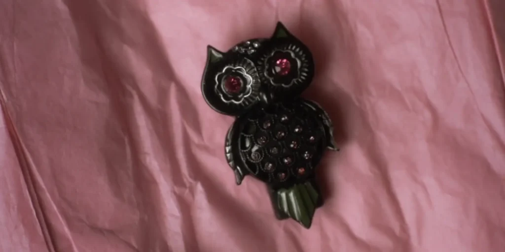 Close up of pendant laying on pink crepe paper. The pendant is a stylized black owl with pink fake jewel eyes and a torso that is studded with smaller pink and silver fake jewels.