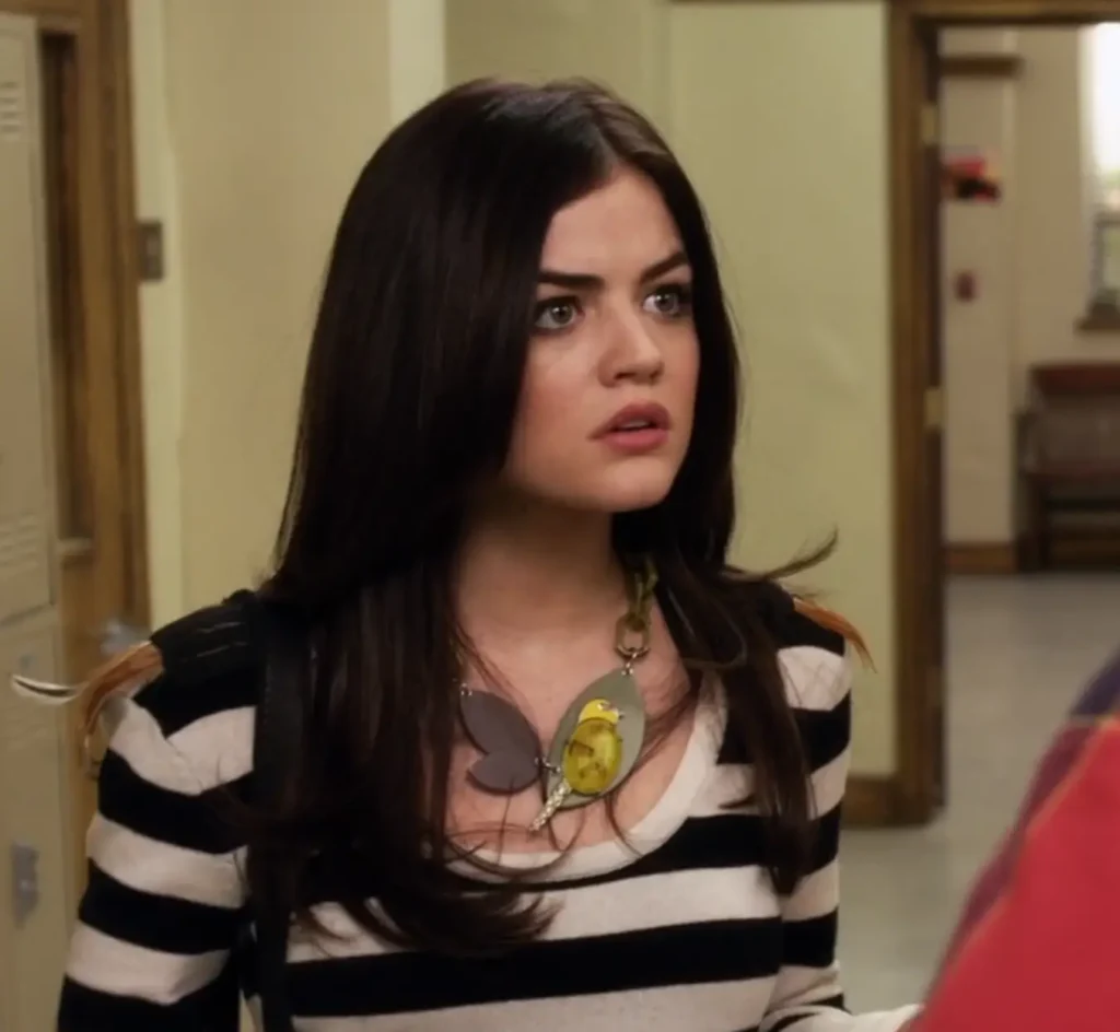 Close-up on Aria, looking concerned. She is wearing a long-sleeved white top with black horizontal stripes and light brown feathers coming off the shoulders. She’s also wearing a large chunky necklace that appears to look like an abstract bird, with a pendant that is a chain with large, visible links.