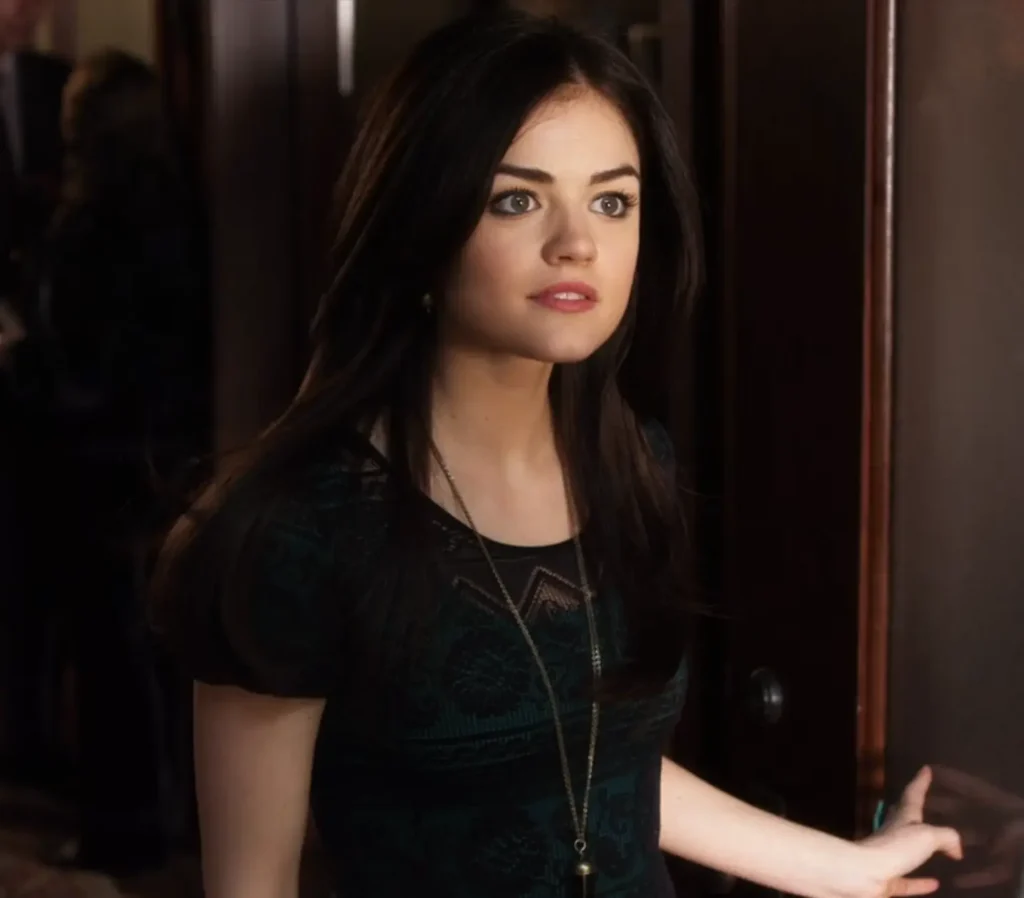 Close-up on Aria answering the door - we can now see that the dress is black and lacy and worn over a dark green shorter dress. She’s also wearing a long necklace with a thin chain and pendant.