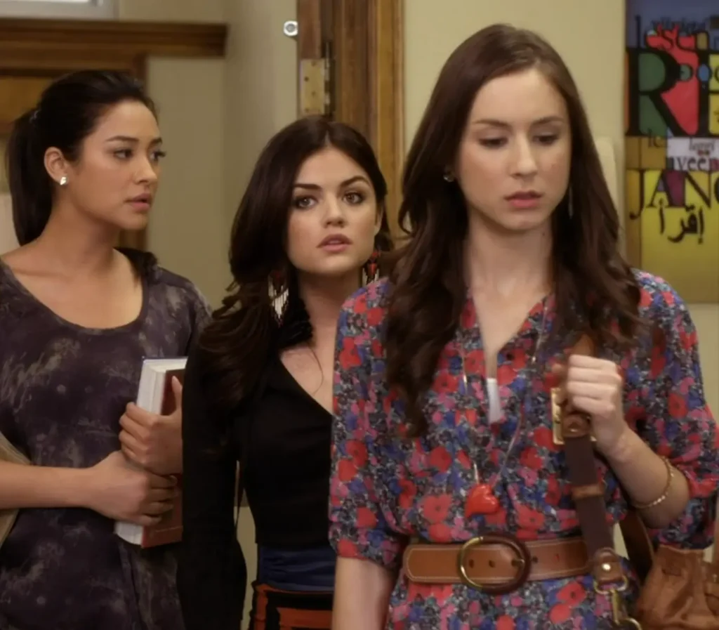 Emily, Aria, and Spencer enter their English classroom. Spencer is wearing a multicolored long button-down with red flowers over a white cami and a brown leather belt over it at the waist, as well as a long thin chain necklace with a large red heart pendant.
