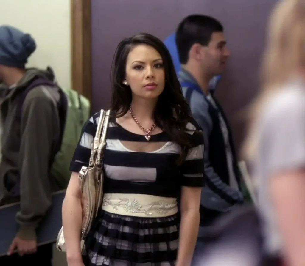 Mona is standing in the school hallway wearing a dress that is transparent with black horizontal stripes, and underneath it is either a white slip or an inner layer of the dress. She is wearing a cream-colored wide belt over it at the waist which has white flowers embroidered on it, and she is wearing a thick copper chain with a small copper heart pendant.