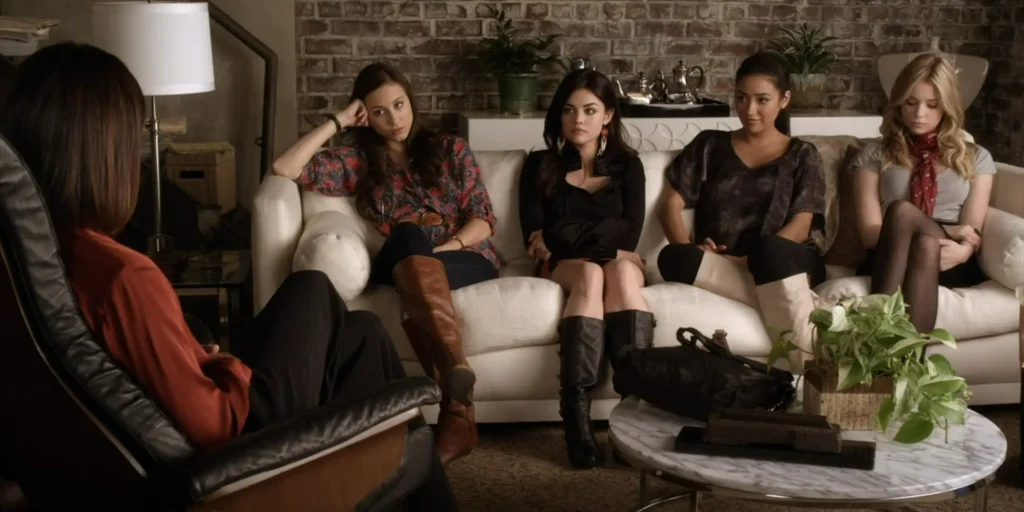 Spencer, Aria, Emily, and Hanna all sit on a couch in Therapist Anne’s office looking sullen. We can now see that Emily’s outfit includes black leggings and knee-high white boots.