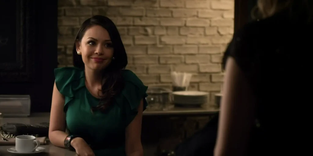 Mona is sitting at a counter at a cafe wearing a dark green top with ruffled short sleeves. She’s smiling at Hanna, who is offscreen.