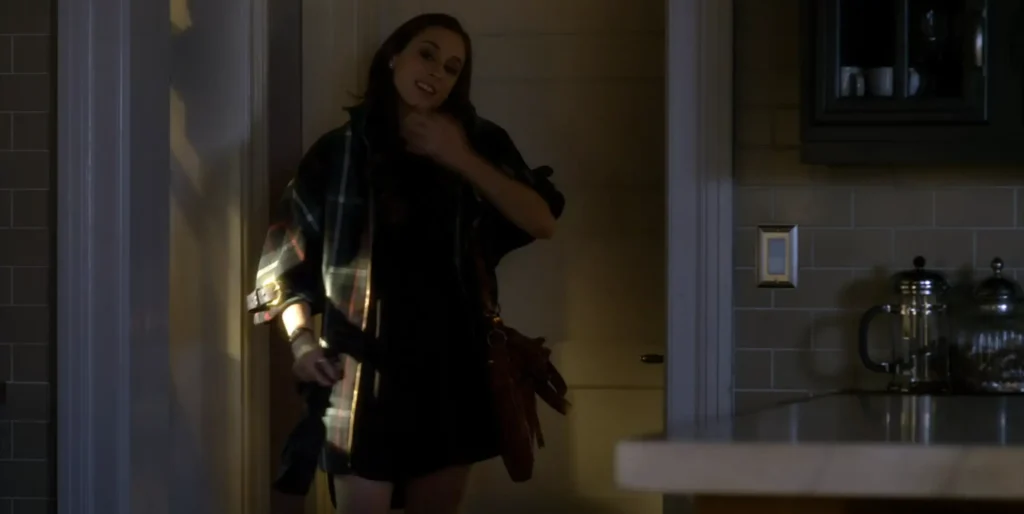Spencer is taking off her coat after just having entered her house; she is mostly in shadow, but we can see that her jacket is plaid in a sort of poncho style.