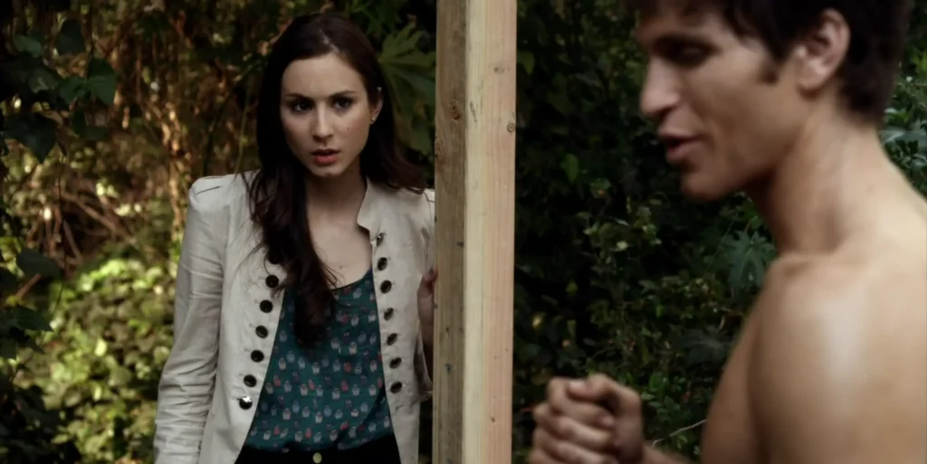 Spencer looks very concerned while watching Toby speak to someone offscreen. Spencer is wearing a blue-green top patterned with tiny owls, tucked into dark jeans, with a white light jacket over it that has large metallic buttons.