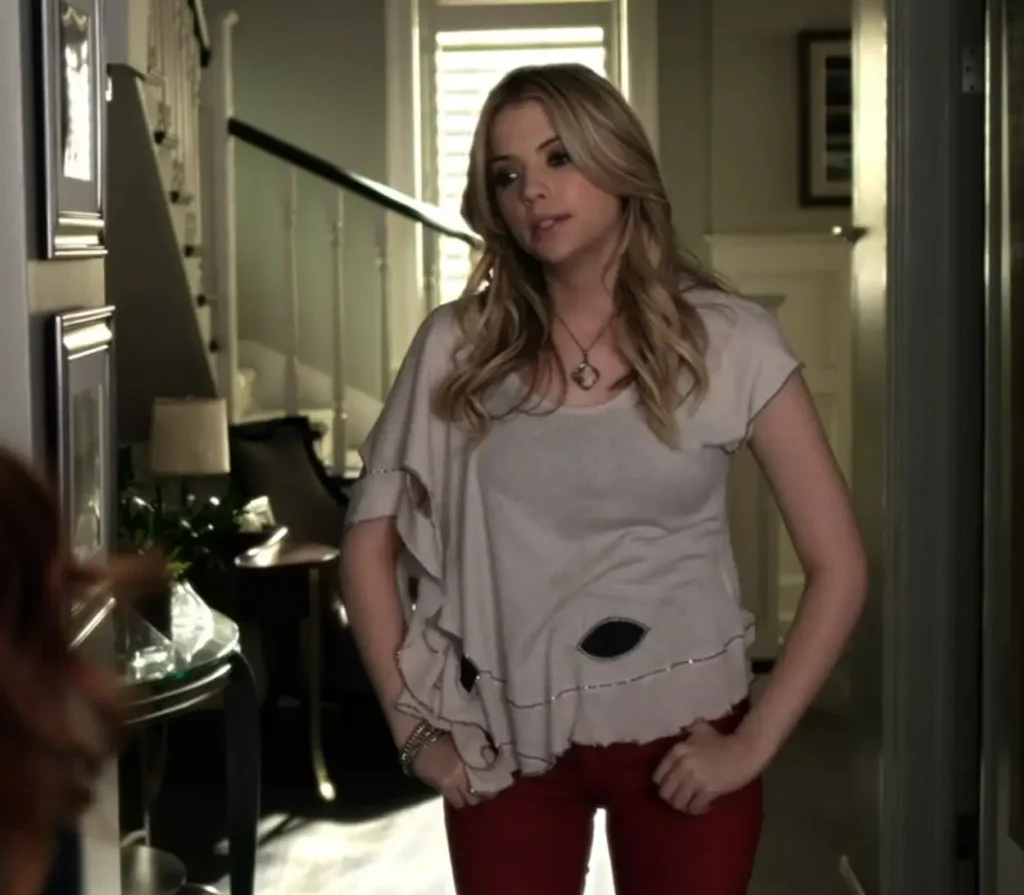 Hanna stands in the hallway of her house talking to Ashley offscreen, looking frustrated with her thumbs hooked in the belt loops of her red jeans. She’s wearing a cream-colored top over a black tank top; the top is assymetrical and has ruffles on one side of the sleeves, as well as largeeye-shaped holes that show the black cami underneath.