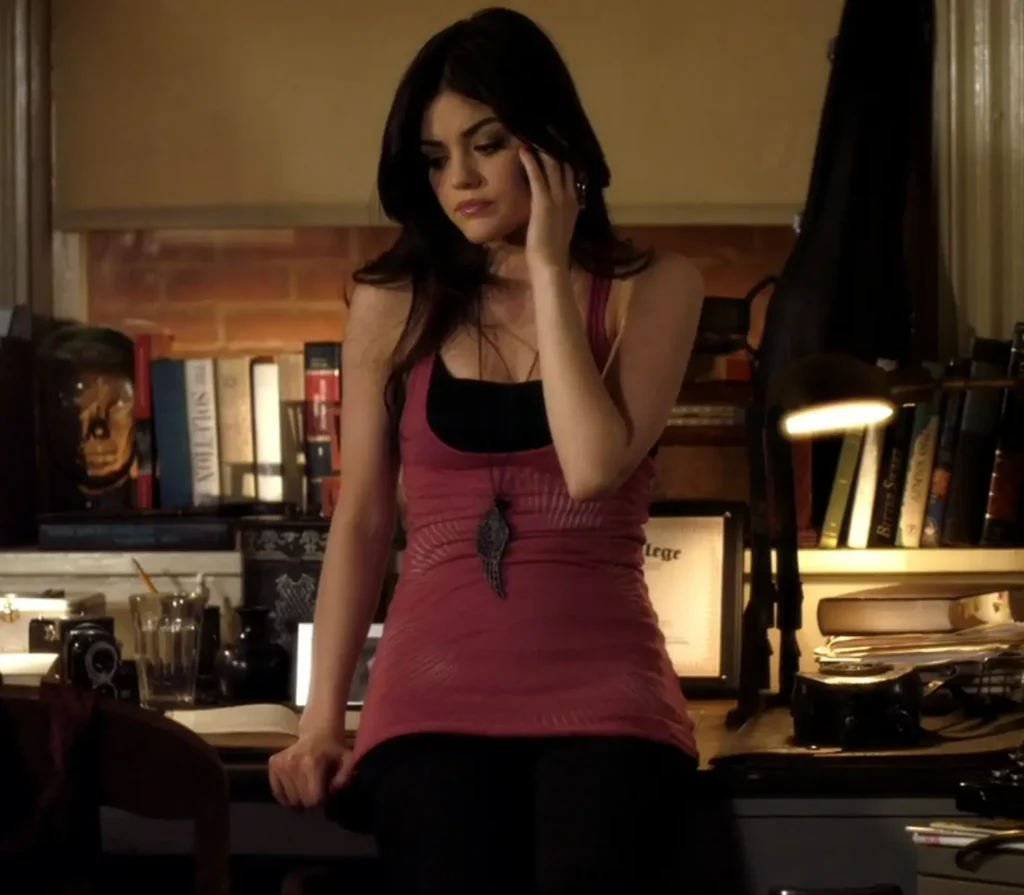 Aria is leaning against a desk, talking on the phone. She’s wearing a long dull red tank top over a black cami and black jeans; the red tank top goes down to her mid-thigh. She also has a necklace that is a thin chain with a very large pendant hanging below her chest.