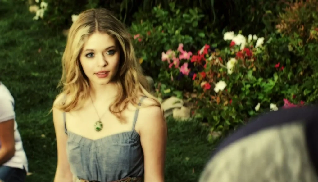 Close up of Ali talking to someone offscreen. We can only see her from the chest up, but she is wearing a light gray sleeveless dress with spaghetti-straps and a necklace