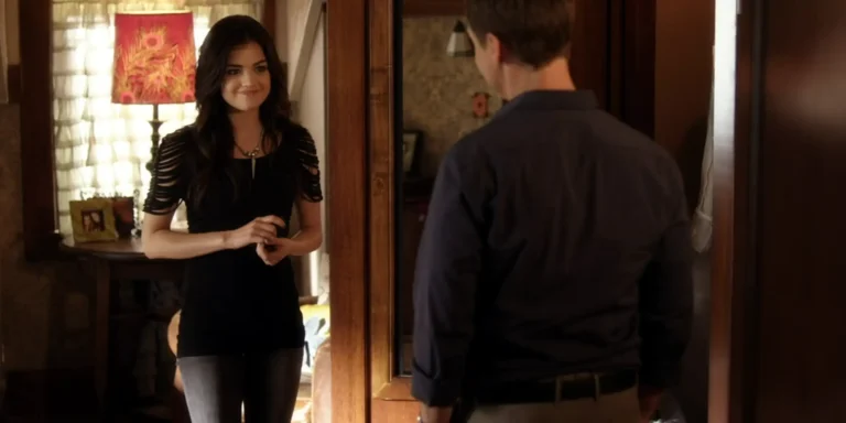 Aria standing in her room smiling at her dad. She’s wearing a black top where the sleeves have narrow strips cut out, and the top goes down to her thigh over distressed gray jeans.