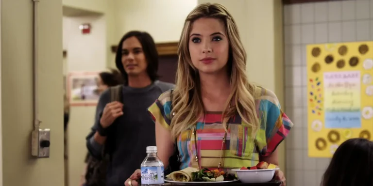 Hanna stands in the Rosewood High cafeteria holding a lunch tray and smiling ruefully while Caleb comes up behind her. Hanna’s wearing a drape-y top with bright multicolored rectangles in an abstract pattern.