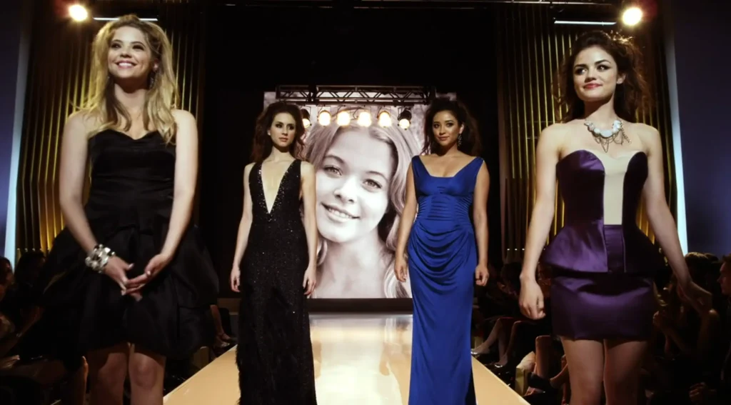 Hanna, Spencer, Emily, and Aria all pose on a fashion runway with a photo of Alison blown up on the screen behind them. Hanna is wearing a black ruffled strapless cocktail dress; Spencer is wearing a full-length sleeveless gown with an extra-deep vee neck and sparkles; Emily is wearing a full-length deep blue sleeveless gown that is bunched up in creases around the middle; and Aria is wearing a dark purple strapless cocktail dress with peplum and a yellow vertical strip down the middle of the cleavage, along with a chunky opal necklace