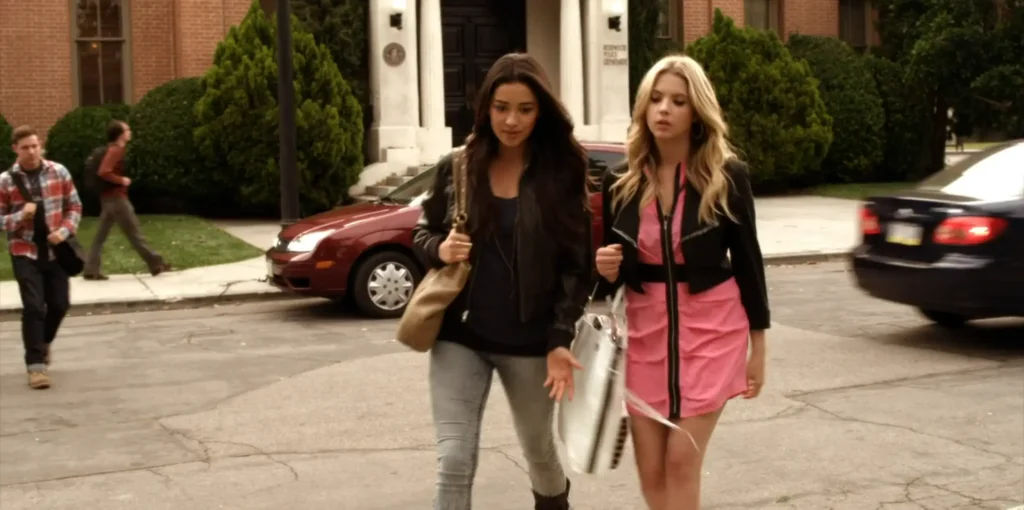 Emily and Hanna walk together towards Rosewood High School. Hanna is wearing a hot pink thigh-length dress with a black zipper running down the middle and a black belt, with a short black jacket on top of it