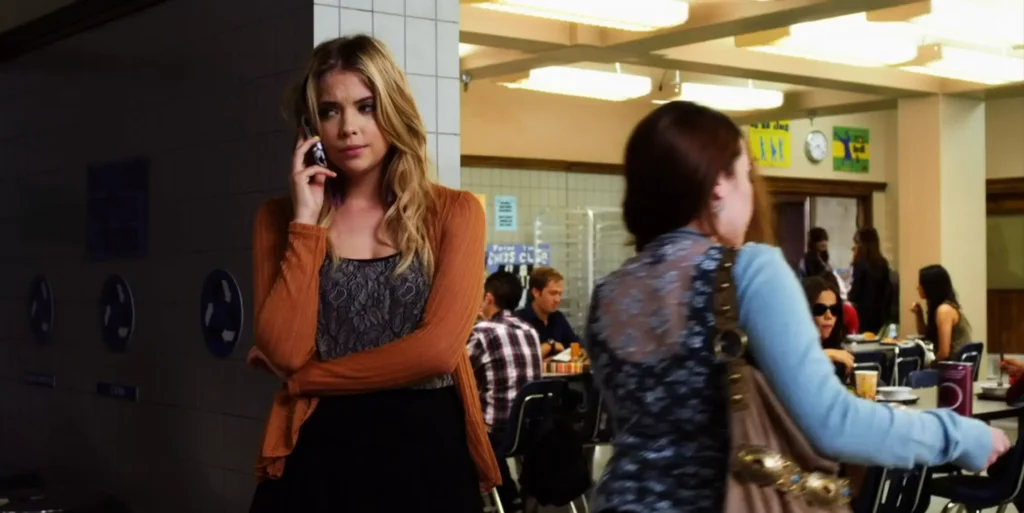 Hanna on the phone in the school cafeteria, looking upset, while wearing a burn orange cardigan over a light gray flower patterned top and a black skirt