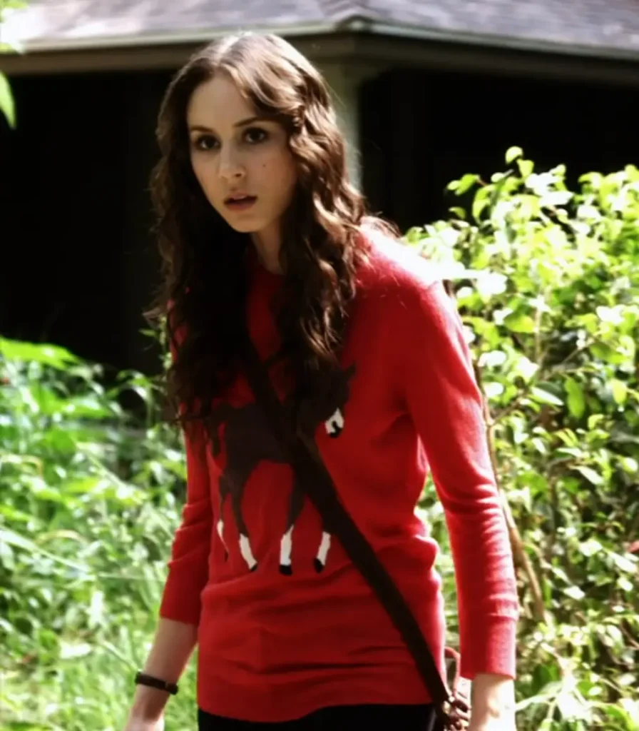 Spencer peering at something with concern while wearing a red sweater with a pattern of a donkey on it, which is partially obscured by her hair and the strap of her bag across her chest