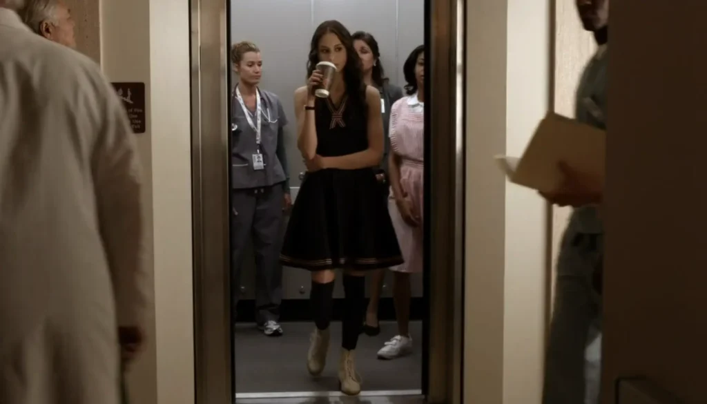 Spencer steps out of an elevator sipping coffee, wearing a knee-length sleeveless navy-blue dress with red trimming that makes it look very sailor-like, with black knee-high socks and short white boots