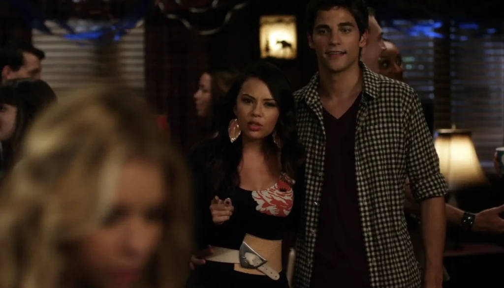 Mona at the party with Noel Kahn. Mona is wearing a dress with a large print of a white and red flower, a wide cream and light brown waist belt, and large red leaf earrings