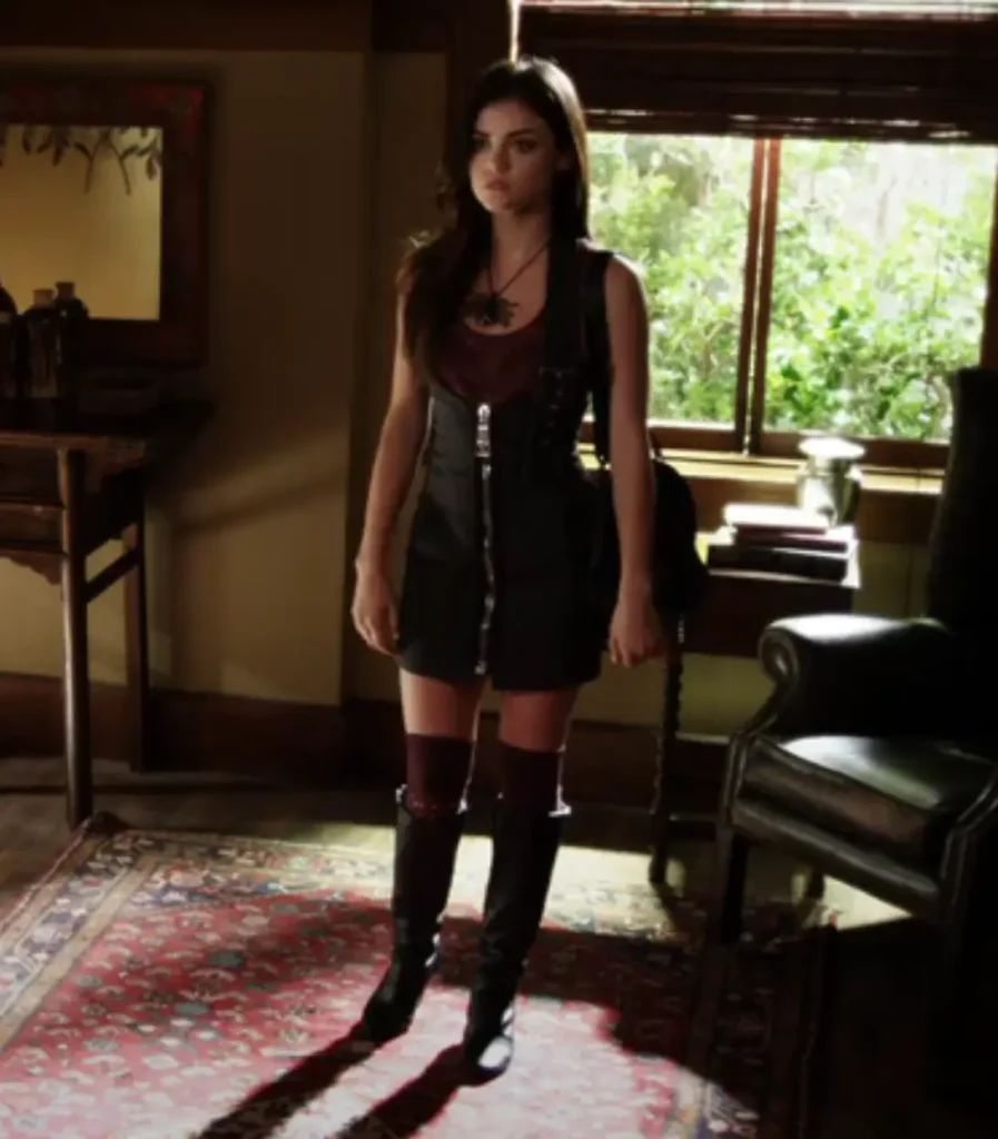 Aria stands in the hallway of her home facing her dad. Aria is wearing a black leather zipper dress over a maroon dark-red top with black leather boots and matching maroon socks. She's also wearing a large black beaded spider necklace.