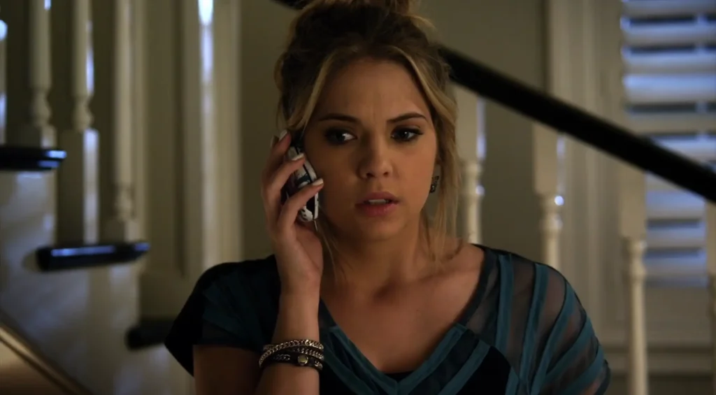 Hanna on the phone looking upset; she is wearing a blue vee-necked blouse with stripes and transparent parts