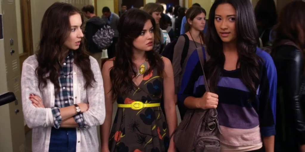 Spencer, Aria, and Emily are walking in the school hallway and talking. Aria is wearing a gray dress with patterns of flowers in coral, black, and dark blue, with a neon yellow waist belt. She also has a necklace with large chunky pendants that may look like a bird