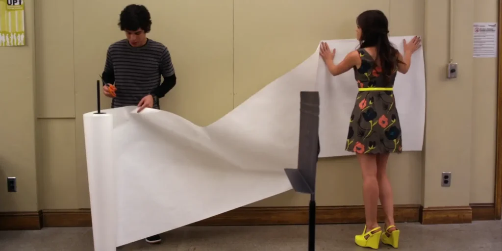Aria and Holden in a classroom putting up a large sheet of paper on the wall. Aria’s back is to the screen, and we can now see that she is wearing neon yellow platform heels that match her belt
