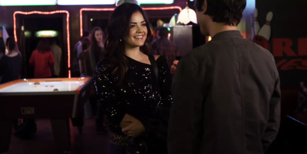 Aria talking to Holden at an arcade; she is smiling brightly and wearing a very sparkly black long-sleeved top with a leopard-print belt visible beneath it