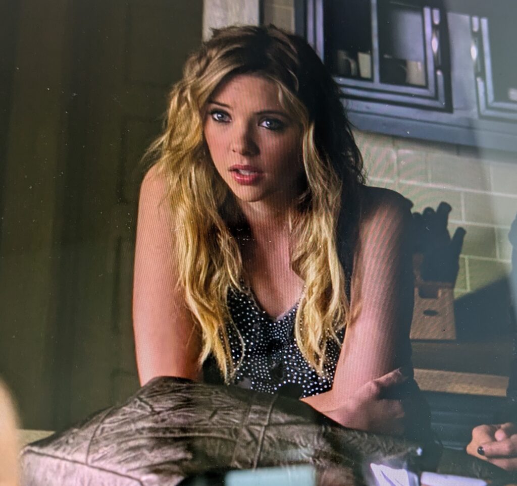 Hanna seated at Spencer’s kitchen counter, leaning forward on her elbows. She is wearing a dark gray sleeveless top with a vee neck and sparkly studs or beads
